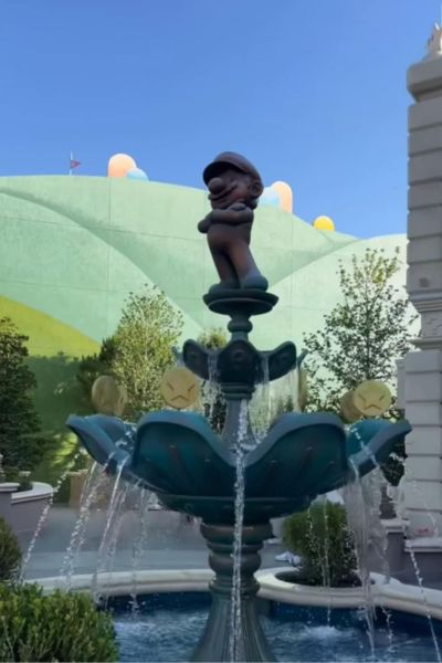A playful statue stands at the top of a water fountain outside with trees in the background.