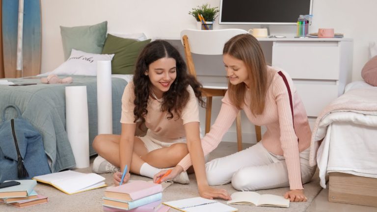 Two college students sitting on the floor in a shared dorm room with beds, books, and study supplies, showing dorm furniture with storage ideas for maximizing a small college space.