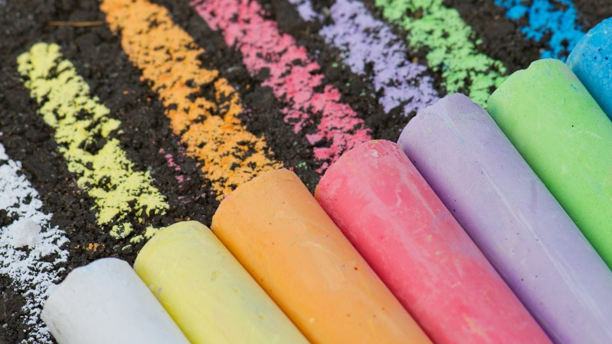 Colorful sidewalk chalk sticks lined up on pavement with bright chalk lines, featuring creative sidewalk chalk drawing ideas for screen free summer fun with kids.